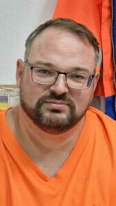 Ein Mann mit Brille und leuchtig orangenem T-Shirt lächelt in Kamera.